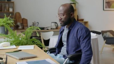 Tekerlekli sandalyedeki Afro-Amerikalı adam oturma odasındaki masaya oturuyor, bilgisayarını açıyor ve uzaktan çalışırken daktilo ediyor.