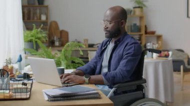 Tekerlekli sandalyedeki Afrikalı Amerikalı adamın portresi masada dizüstü bilgisayarda çalışıyor ve sonra evdeki oturma odasında kameraya poz veriyor.