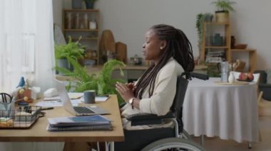 Tekerlekli sandalyedeki Afro-Amerikalı kadın, iş günü e-toplantısı sırasında laptoptan çevrimiçi video görüşmesi yaparak konuşuyor.