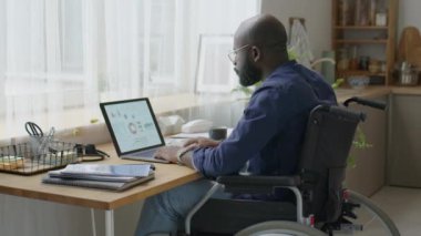 Tekerlekli sandalyedeki Afrikalı Amerikalı işadamı ev işyerindeki laptopta finansal verilerle çalışıyor.