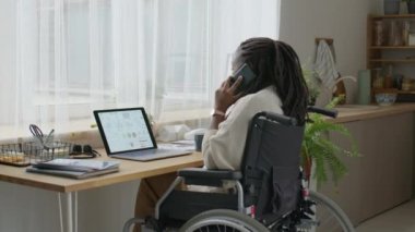 Tekerlekli sandalyedeki Afrikalı Amerikalı iş kadını telefonda konuşuyor ve evde çalışırken bilgisayardaki finansal verileri analiz ediyor.