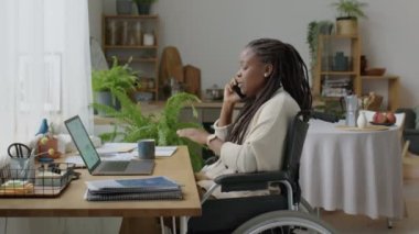 Tekerlekli sandalyedeki Afro-Amerikalı kadının telefon görüşmeleri ve uzaktan çalışırken bilgisayarını kullanışının yan görüntüsü.