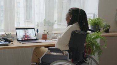 Arka planda, tekerlekli sandalyede oturan ve evinde uzaktan çalışırken bilgisayardan iş arkadaşlarıyla konuşan Afrikalı Amerikalı bir kadın var.