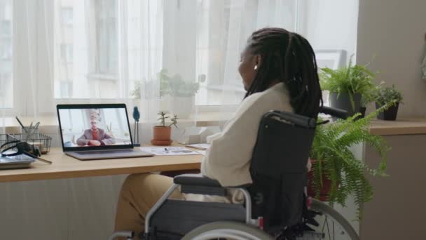 アフリカ系アメリカ人女性が机の車椅子に座り 自宅で遠隔操作しながらノートパソコンで同僚と話す様子 — ストック動画