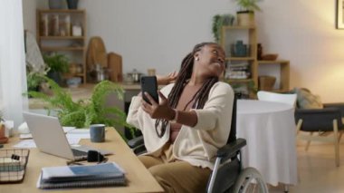 Küçük oğluyla evde selfie çekerken tekerlekli sandalyede gülümseyen ve akıllı telefon kamerasına poz veren Afrikalı Amerikalı bir kadın.