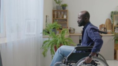 Evdeki oturma odasında tekerlekli sandalyede oturan siyah bir adam, gözlüğünü çıkarıyor, gözlerini ovalıyor ve pencereden bakıyor.