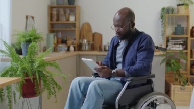 Engelli Afrikalı Amerikalı bir adamın tekerlekli sandalyede oturup dijital tablet kullandığı orta boy bir fotoğraf.