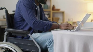 Tekerlekli sandalyedeki siyahi adamın meyve suyu içip evdeki masada dizüstü bilgisayarda daktilo kullandığı sahneler.