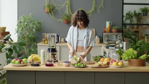 stock video Medium long shot of young woman cutting fresh green apple at kitchen table while making fruit smoothie at home