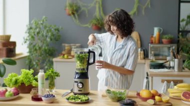 Genç bir kadının evdeki mutfakta yeşil smoothie yaparken miksere su doldurduğu orta boy bir fotoğraf.