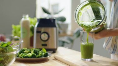 Bardakları evdeki mutfaktaki blenderdan yeni yapılmış yeşil meyve püresiyle dolduran bir kadının seçici görüntüsü.