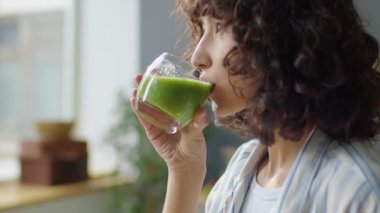 Genç kızın camdan yeni yapılmış yeşil smoothie 'yi içerken ve evde dikilirken pencereden bakarken orta boy yakın çekimi.