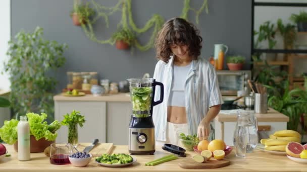自宅でキッチンで緑のスムージーを調理しながら 新鮮なサラダの葉をブレンダーに置く若い女性 — ストック動画