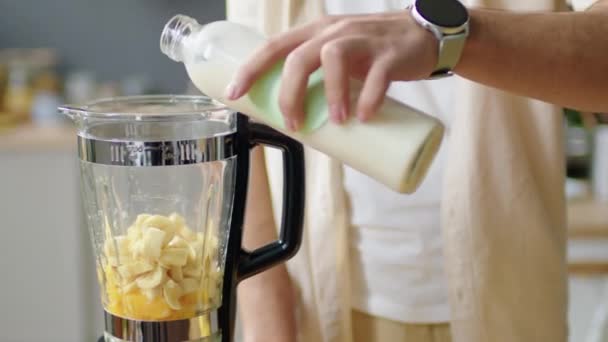 自宅でキッチンでスムージーを作っている間 ブレンダーと閉じたふたで新鮮な果物にビーガンミルクを追加する男性のクロップされたインペラントショット — ストック動画