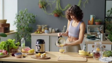 Spor sütyenli ve tozluklu genç bir kadının blenderdan cama meyve suyu dökerken ve mutfakta içerken orta uzunlukta bir pozu.