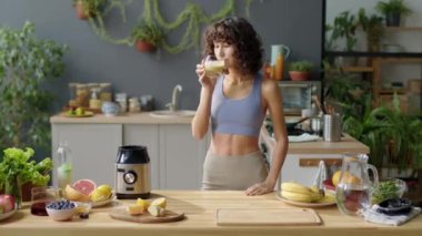 Orta boy spor giyinen, camdan meyve kokteyli içen ve evde gülümseyerek kameraya poz veren seksi bir kız.