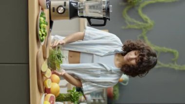 Mutfak masasında gülümseyen ve kameraya poz veren vejetaryen kızın dikey portresi. Üstünde taze meyve, sebze ve blender var.