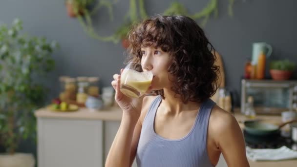 在厨房里 穿着运动服的漂亮姑娘用玻璃杯轻饮水果 在镜头前微笑着摆出一副中等特写 — 图库视频影像