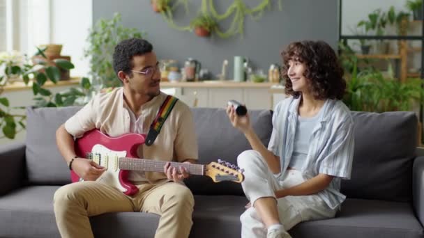 Junger Mann Sitzt Auf Sofa Wohnzimmer Spielt Gitarre Und Shaker — Stockvideo