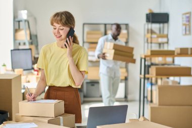 Deponun genç ve gülümseyen bayan müdürü cep telefonuyla müşteriyle konuşuyor ve paketlenmiş kutunun üstüne kişisel bilgileri yazıyor.
