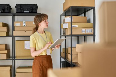 Tabletli genç bir bayan yönetici, içinde sipariş edilmiş mallar ve kontrol bilgileri olan karton kutular ile uzun rafların önünde duruyor.