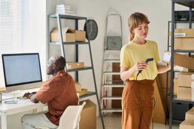 Tarayıcı ile genç bir kadın paketlenmiş karton kutulardaki kodları kontrol ediyor. İçinde bilgisayarlı erkek meslektaşlarına karşı sipariş edilmiş eşyalar var.
