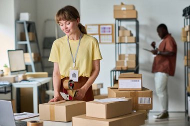 Sarı tişörtlü genç bir kadın iş yerinde kameranın önünde dururken seloteyp ile karton kutuyu mühürlüyor.