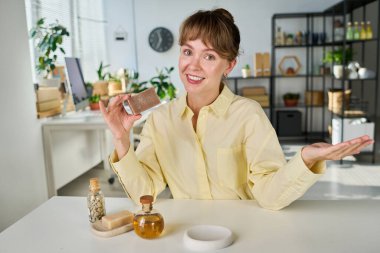 Gülümseyen genç kadın yeni el yapımı sabun ve kendi yarattığı diğer kişisel aşk ürünlerini tanıtırken kameranın karşısında oturuyor.