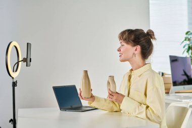 Çevrimiçi izleyicilere yeni vazo koleksiyonundan bahseden iki el yapımı vazoyla gülümseyen genç bir kadın.
