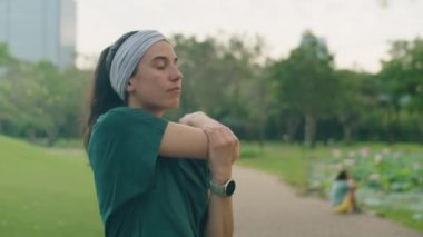 Spor kıyafetli genç bir kadının parkta antrenmandan önce kollarını, bileklerini ve omuzlarını esneterek ısınırken orta kavisli pozu.