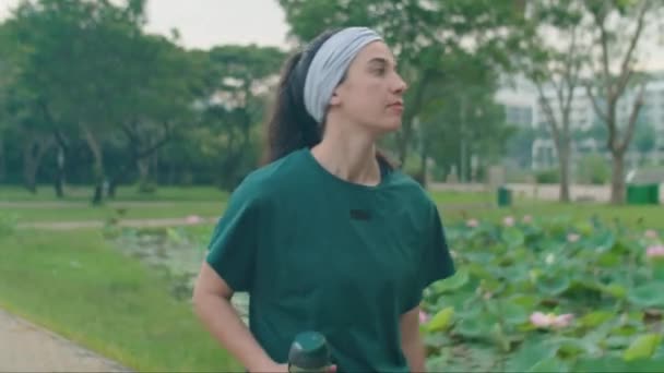 一名身穿运动服的年轻女子拿着水瓶 在公园散步进行晨跑时检查时间的中景照片 — 图库视频影像