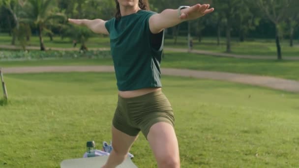 穿着运动服的年轻女子在公园的绿地草坪上练习瑜伽时 在垫子上摆出一副战士的姿势 — 图库视频影像