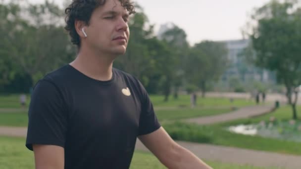 在公园里 穿着运动服 手持无线耳机 头戴莲花的男人 在早晨瑜伽时 他们坐在垫子上 闭眼沉思 手握泥巴 — 图库视频影像