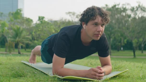 在公园进行户外运动时 身穿运动服和带无线耳机的男子在健身垫上做跳板运动 — 图库视频影像