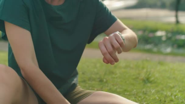 スポーツウェアとヘッドバンドに座っている若い裸足の女性のショットをキルトアップ エクササイズマン ウォーターボトルを保持し 公園で屋外のトレーニングの後に休んでいる間 スマートウォッチを使用して — ストック動画