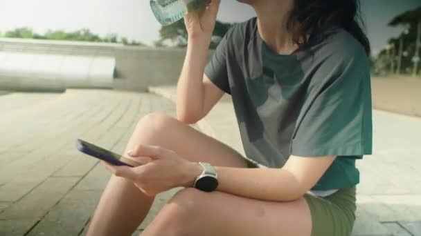 在城市里 穿着运动服和头饰坐在户外 用瓶子喝水 早上跑步后休息时使用智能手机的年轻女子的倒立镜头 — 图库视频影像