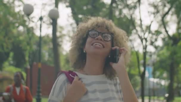バックパック笑顔でアジアのカレッジガールのミディアムクローズアップショットと勉強後に公園を歩いている間の電話でチャット — ストック動画