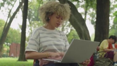 Kıvırcık saçlı Latin üniversiteli bir kızın parkta oturup dizüstü bilgisayarında yazı yazmasının düşük açılı görüntüsü.