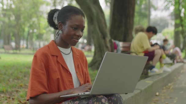 非洲裔美国女大学生坐在停车场路边 一边笑着一边用笔记本电脑上网的中景照片 — 图库视频影像