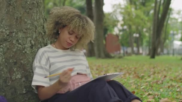 Medium Laukaus Latinalaisen Naispuolinen Opiskelija Istuu Puussa Puistossa Tekee Muistiinpanoja — kuvapankkivideo