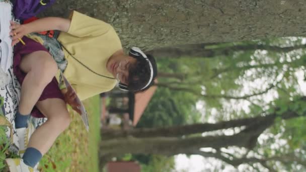 描述亚洲男生坐在公园里一棵树旁 一边看书 一边在剪贴板上记笔记的耳机中的垂直照片 — 图库视频影像