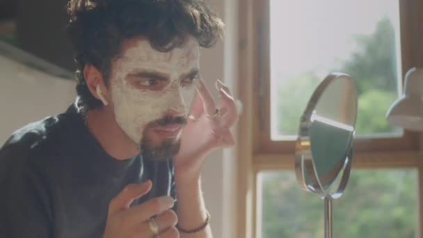 ワイヤレスイヤホンの若者のミディアムクローズアップショットは 自宅でタブレットミラーの前に顔に粘土マスクを適用しながら音楽を聞きます — ストック動画
