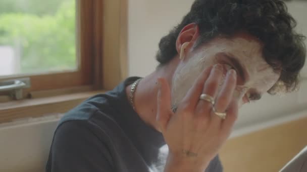 自宅でタブレットミラーの前で顔のマスクを適用しながら 音楽を聞いているワイヤレスイヤホンの若者のミディアムクローズアップショット — ストック動画