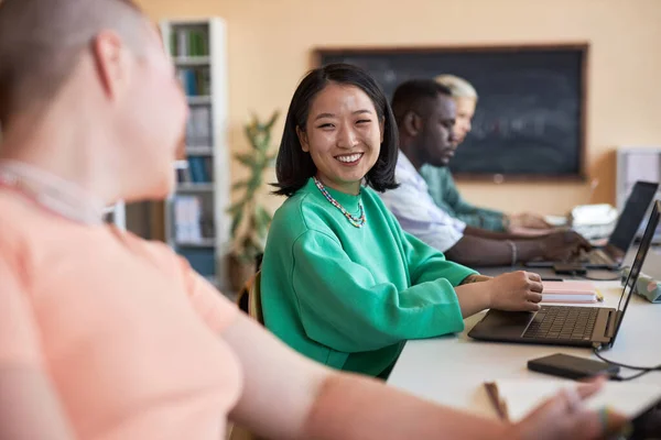 年轻而快乐的亚洲女人坐在笔记本电脑前的桌子旁 看着她的同学或同事 讨论工作要点 — 图库照片