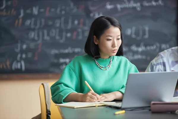 身穿绿色套头衫的年轻沉思女学生一边看笔记本电脑屏幕 一边分析在线数据 并在课上抄本上记笔记 — 图库照片