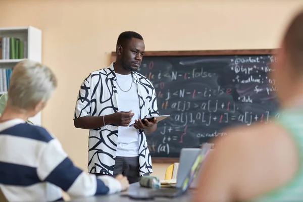 拿着平板电脑的非裔美国人一边站在黑板旁边一边一边听着同学们在研讨会或计算机科学课上的演讲 一边拿着公式 — 图库照片