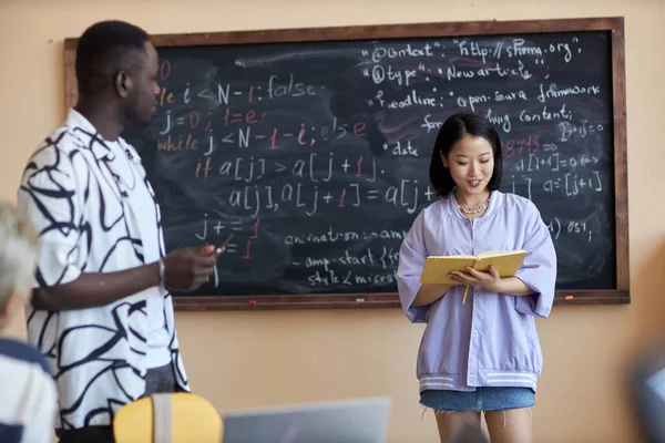 亚洲女学生一边站在黑板旁边 一边看着打开的书 一边拿着公式在同学面前做报告 — 图库照片