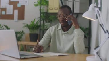 Ofiste çalışırken Afrikalı Amerikalı iş adamının cep telefonuyla konuşup not aldığı orta boy bir fotoğraf.