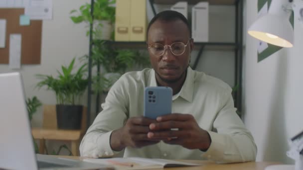 アフリカ系アメリカ人のオフィスワーカーがデスクに座りながらスマートフォンでインターネットを閲覧し笑顔で撮影 — ストック動画