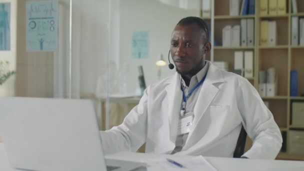 Średnie Ujęcie Czarnego Lekarza Słuchawkach Białym Płaszczu Siedzącego Przy Biurku — Wideo stockowe